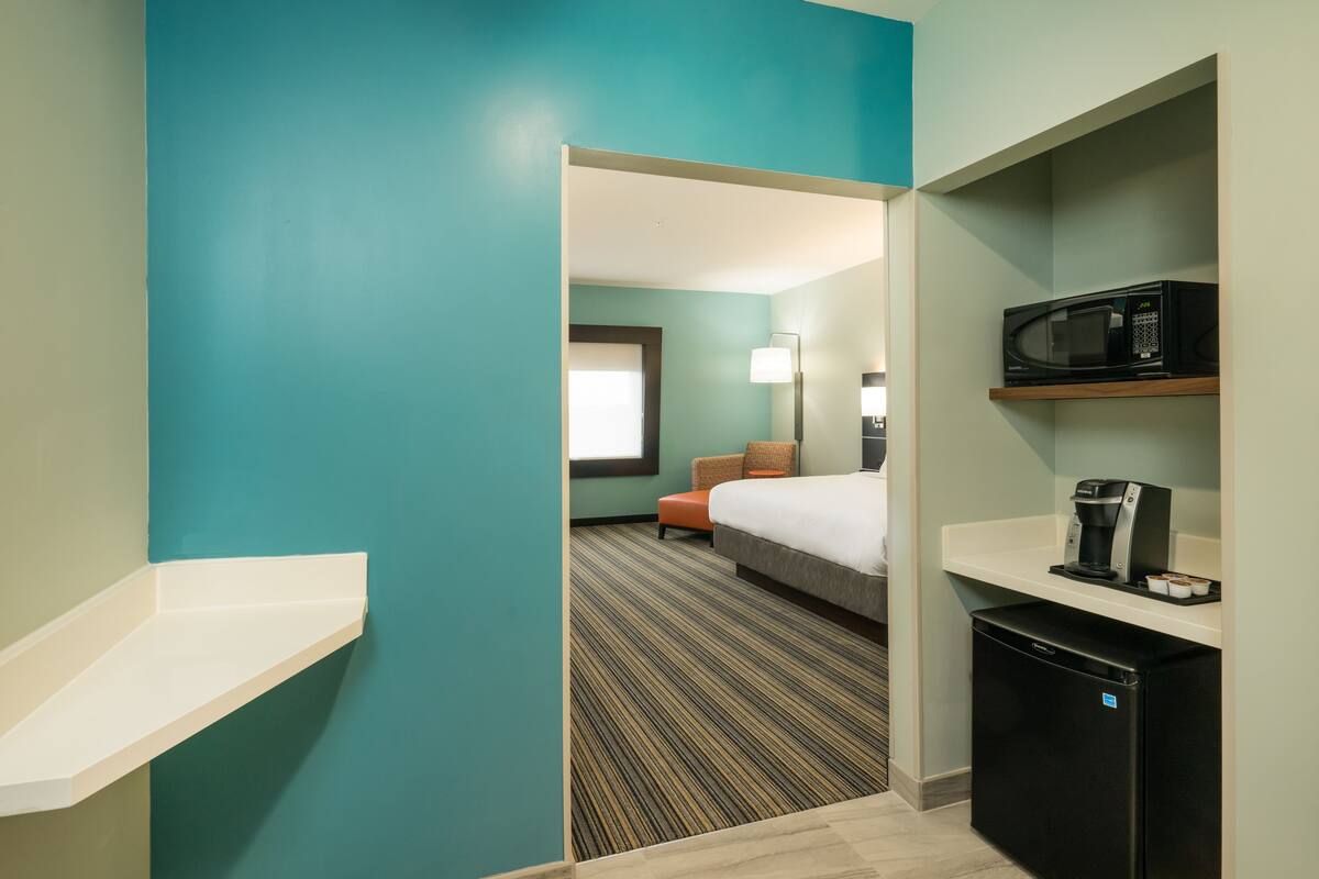 Ein Hotelzimmer mit türkisfarbenen Wänden, einem Bett, einem Schreibtisch mit Mikrowelle, einer Kaffeemaschine und einem kleinen Kühlschrank.