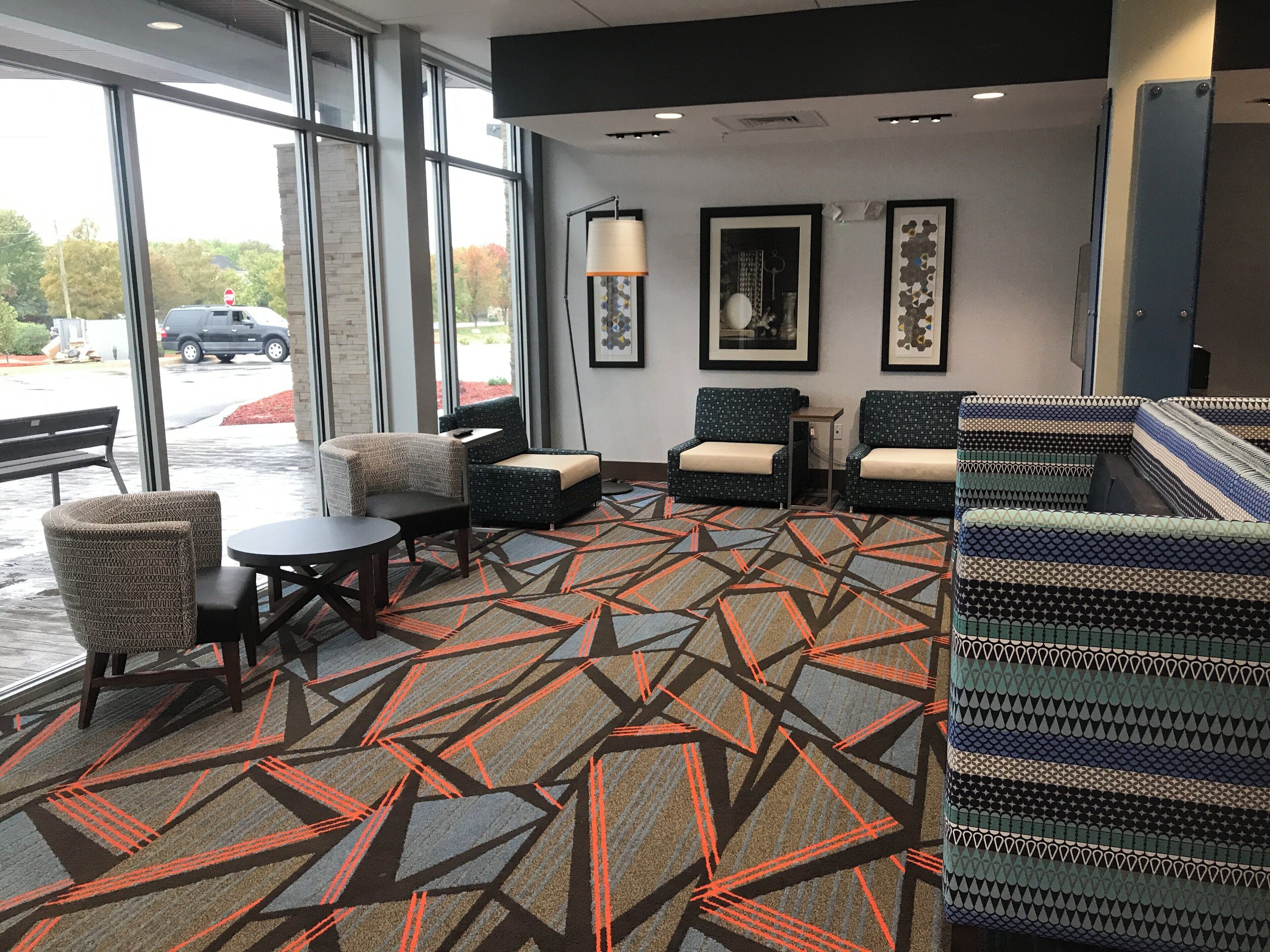 A hotel lobby with a patterned carpet, seating area with chairs and a table, framed artwork on the walls, and large windows.