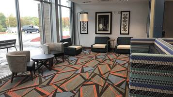 A hotel lobby with a patterned carpet, seating area with chairs and a table, framed artwork on the walls, and large windows.