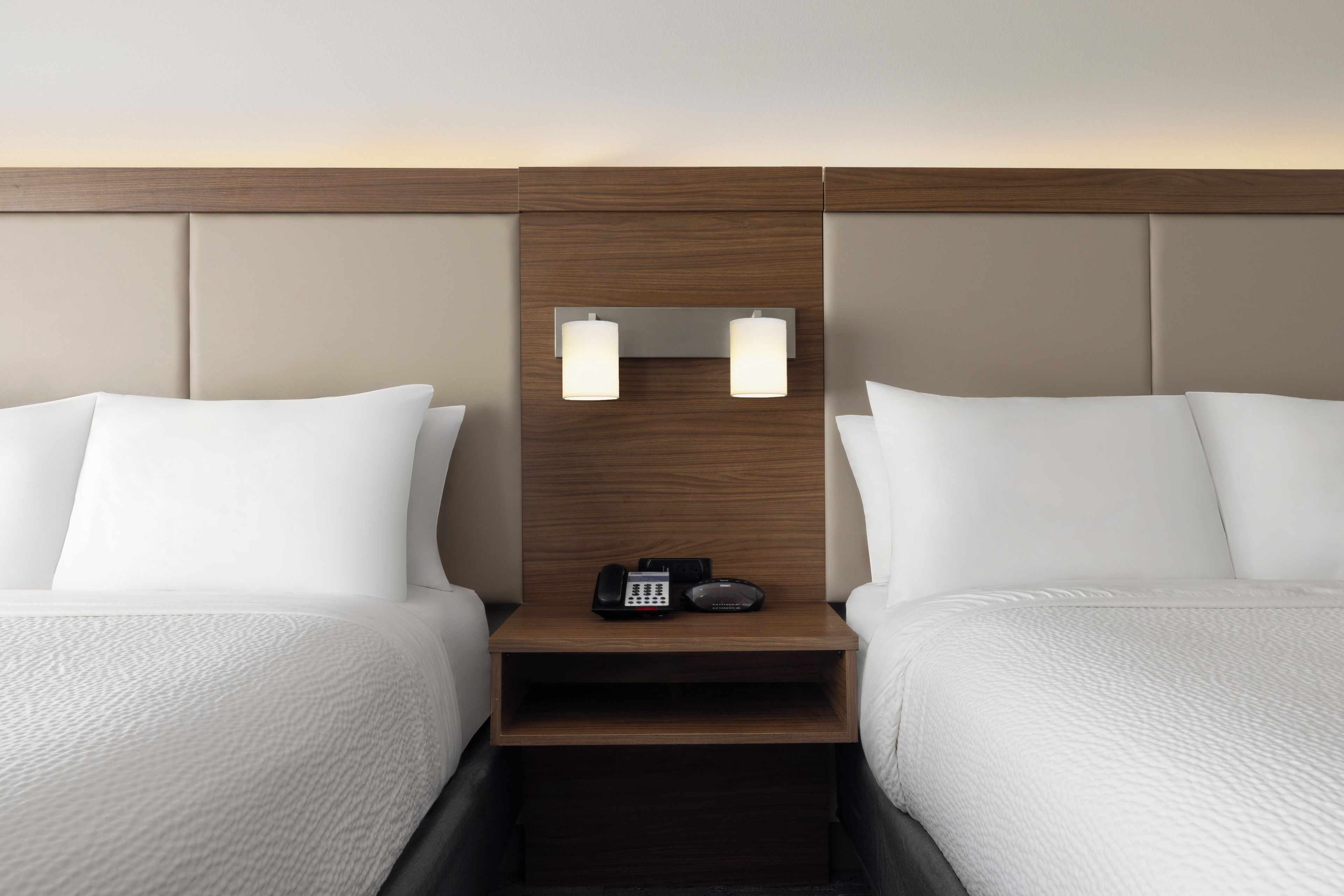 A hotel room with two beds, a wooden headboard, a nightstand with a phone, and wall-mounted lights.