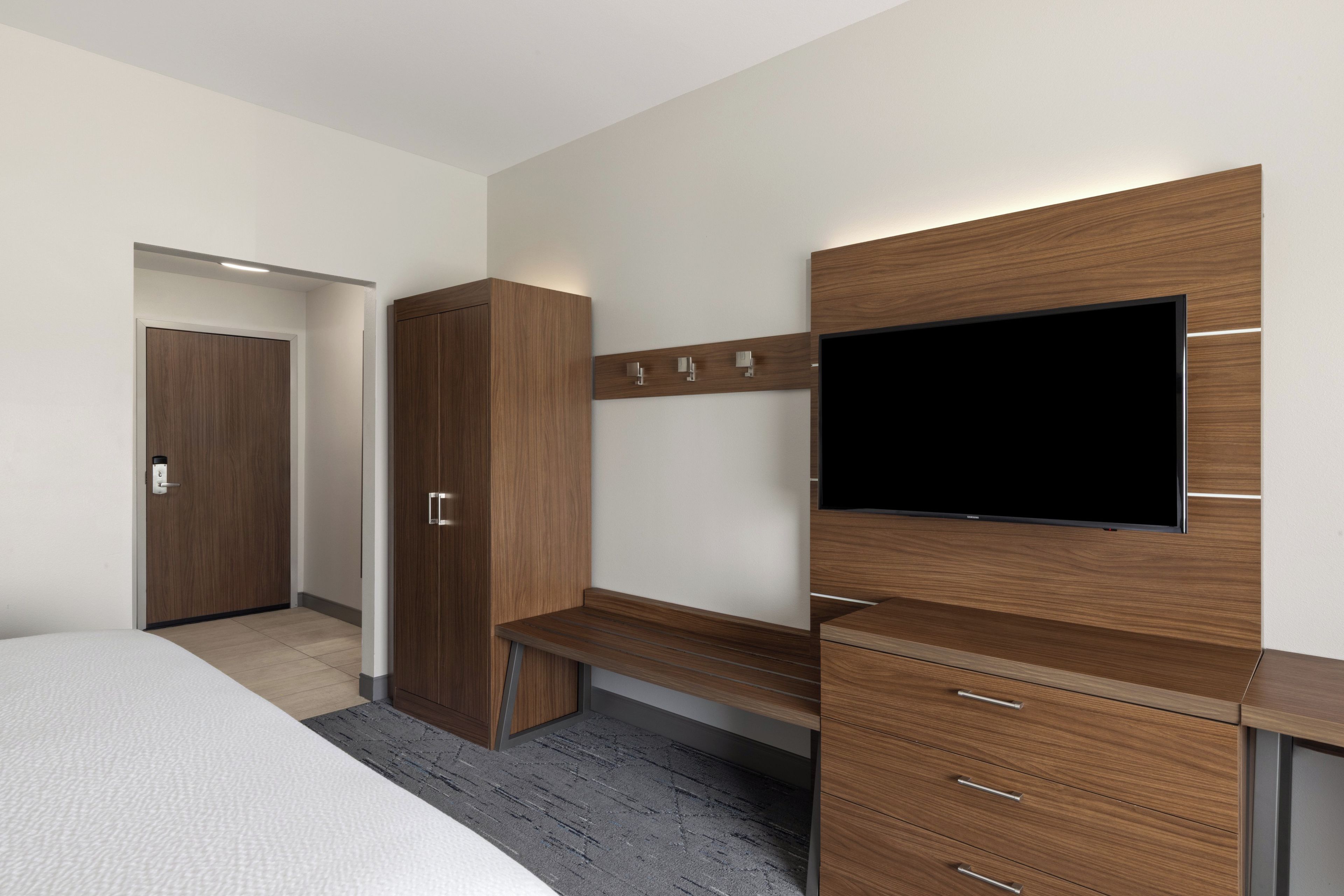 A modern hotel room with a bed, a wooden wardrobe, a desk with drawers, a flat-screen TV mounted on the wall, and a door leading to another room.