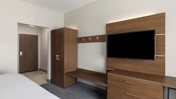 A modern hotel room with a bed, a wooden wardrobe, a desk with drawers, a flat-screen TV mounted on the wall, and a door leading to another room.