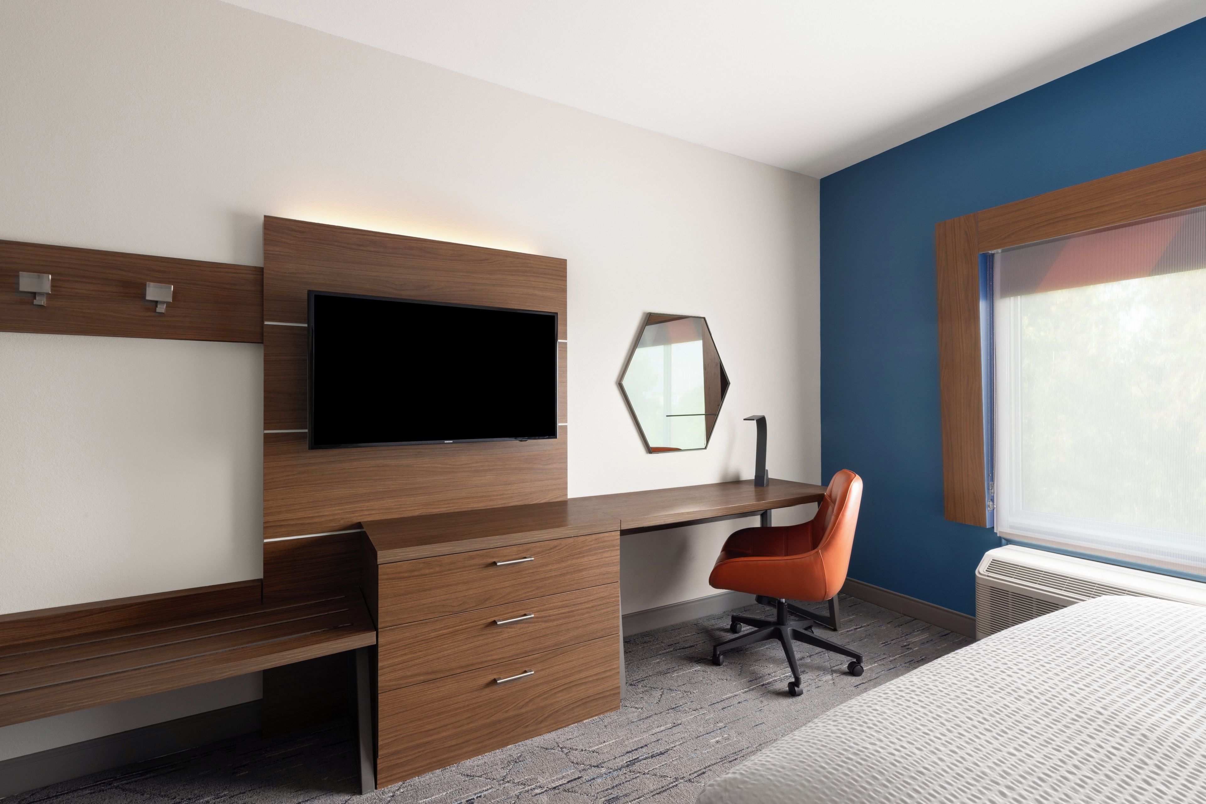 A modern hotel room with a wooden desk, a flat-screen TV, a mirror, a single orange chair, and a large window.