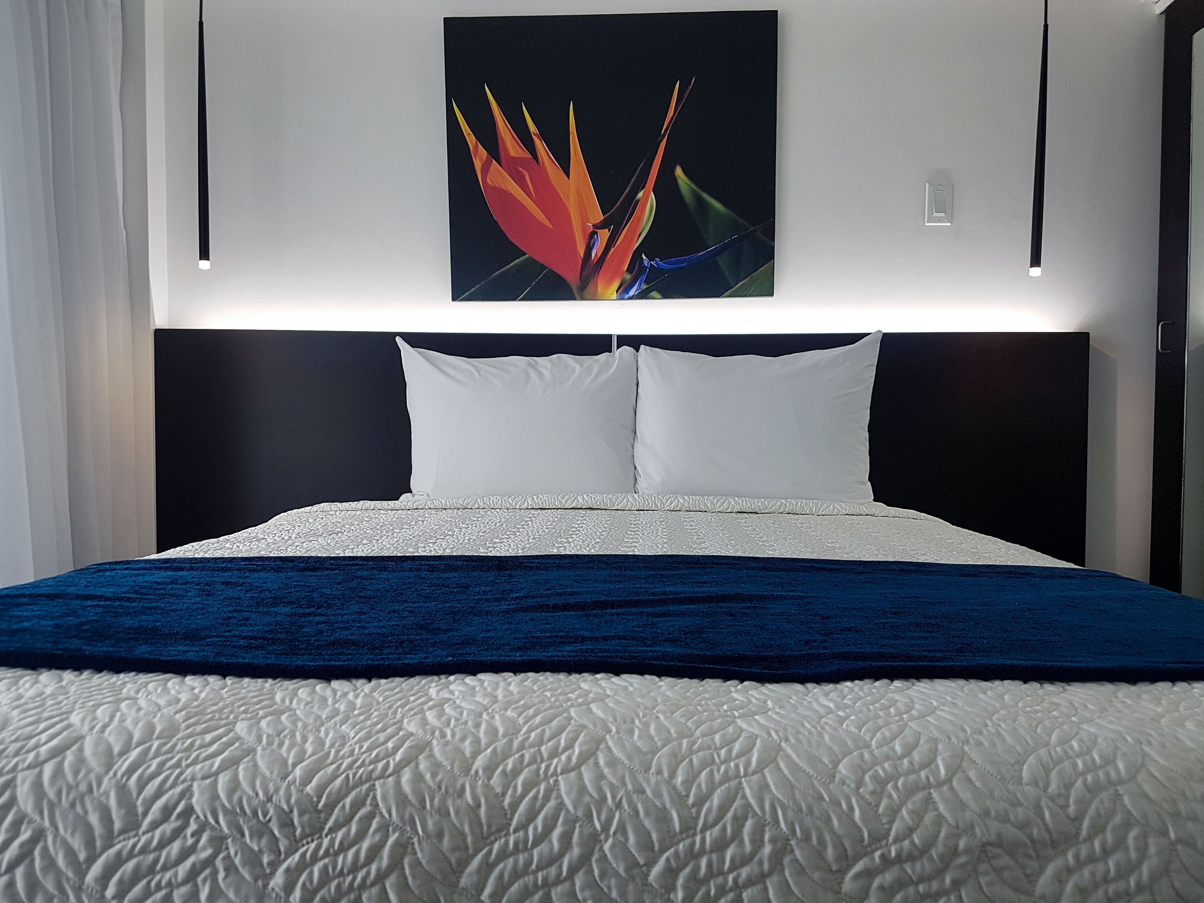 A hotel room with a large bed, two pillows, a dark headboard, a wall-mounted light, and a framed picture above the bed.