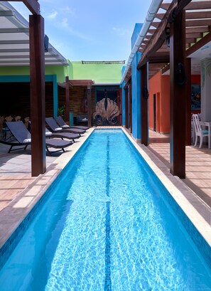 A swimming pool with lounge chairs and umbrellas, surrounded by wooden pillars and a covered area with a green roof.