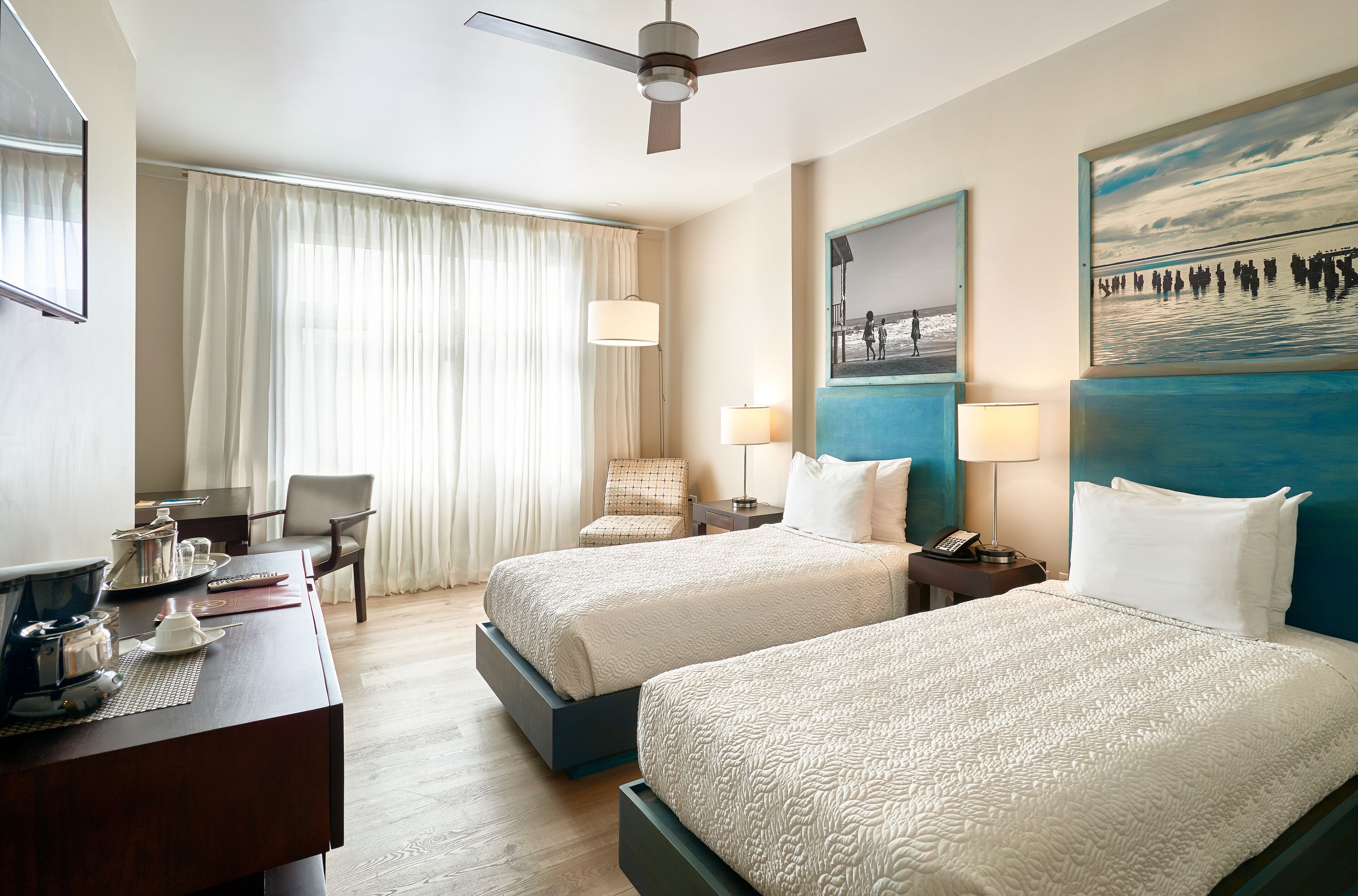 A hotel room with two beds, a desk with a coffee maker, a chair, and a large window with curtains.