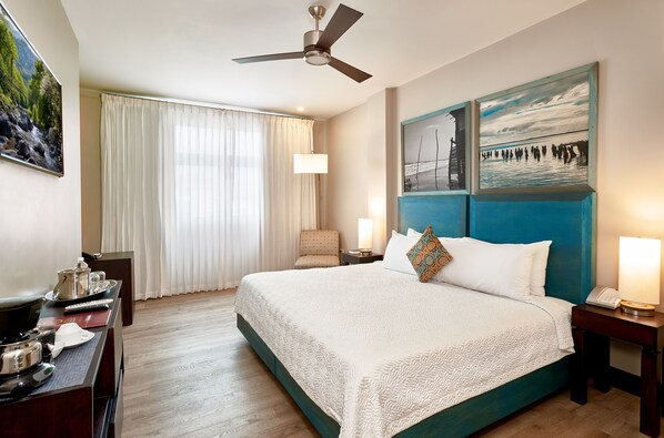 A hotel room with a large bed, a ceiling fan, a desk with a lamp, and a view of the outdoors through the window.