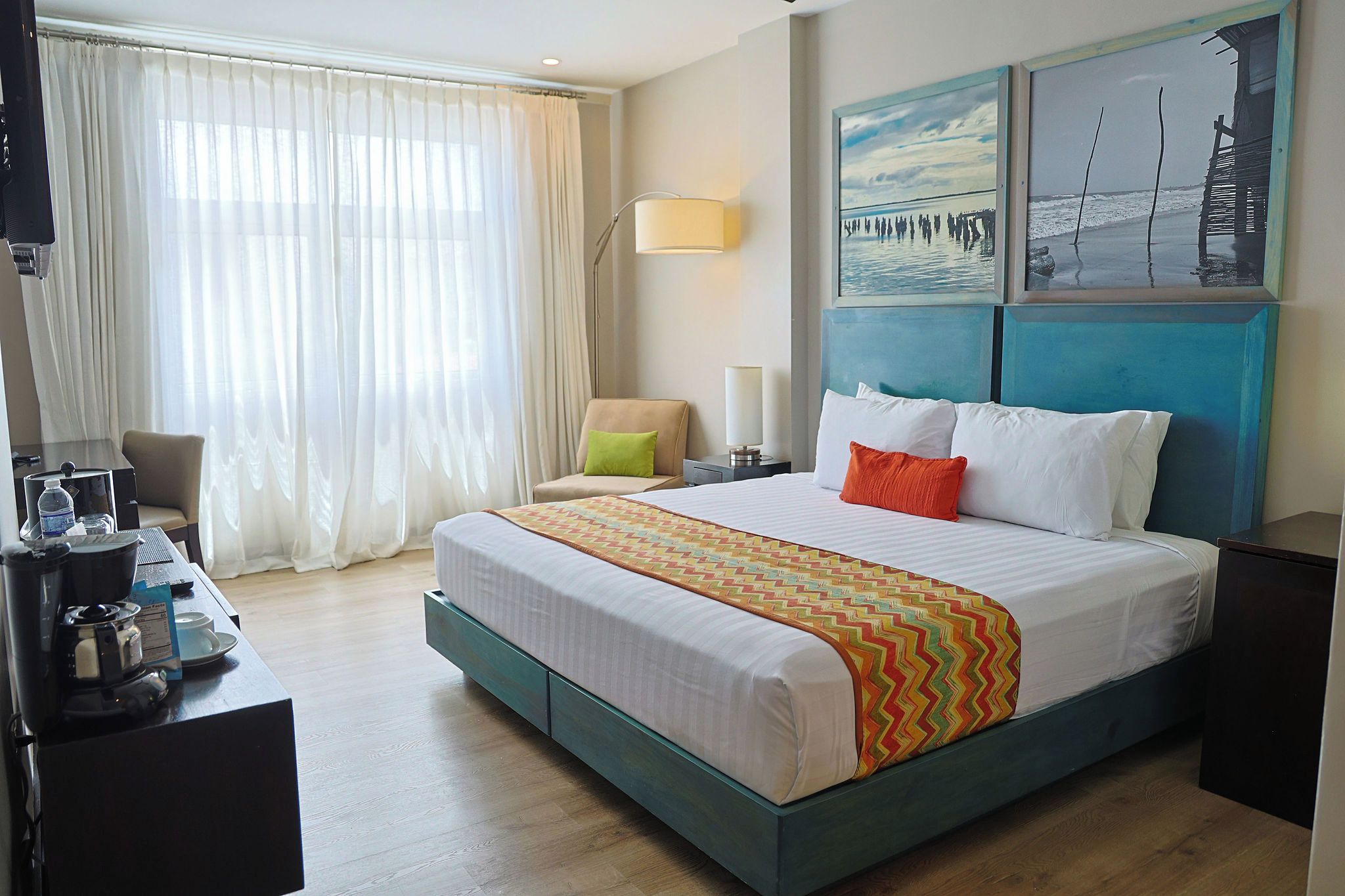 A hotel room with a bed, a desk with a coffee maker, a chair, a lamp, and artwork on the wall.
