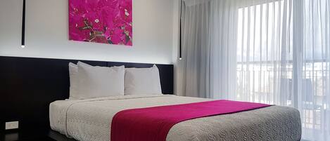 Ein modernes Hotelzimmer mit einem großen Bett, weißer Bettwäsche, einer roten Tagesdecke und einem abstrakten Gemälde in Rosa an der Wand.
