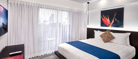 Una habitación de hotel moderna con una cama grande, cortinas blancas, un televisor montado y un cuadro en la pared.