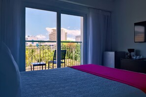 A hotel room with a bed, a desk, a chair, and a balcony with a view of buildings and greenery.