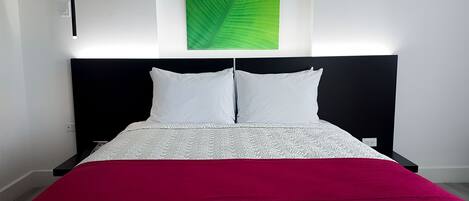 Cama bem arrumada com colcha branca e vermelha, obra de arte com folha verde na parede e cabeceira preta.
