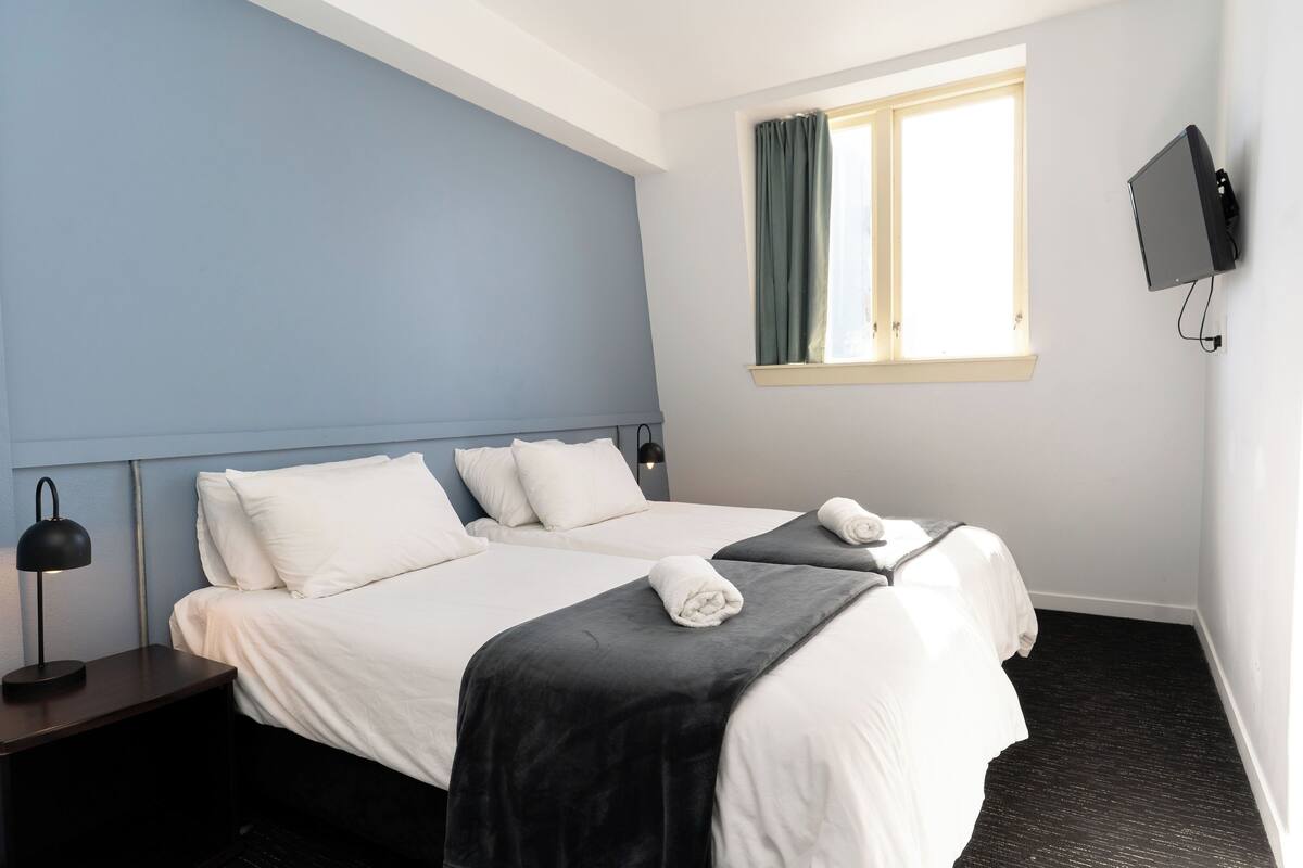 A hotel room with two beds, a TV mounted on the wall, and a window with curtains.