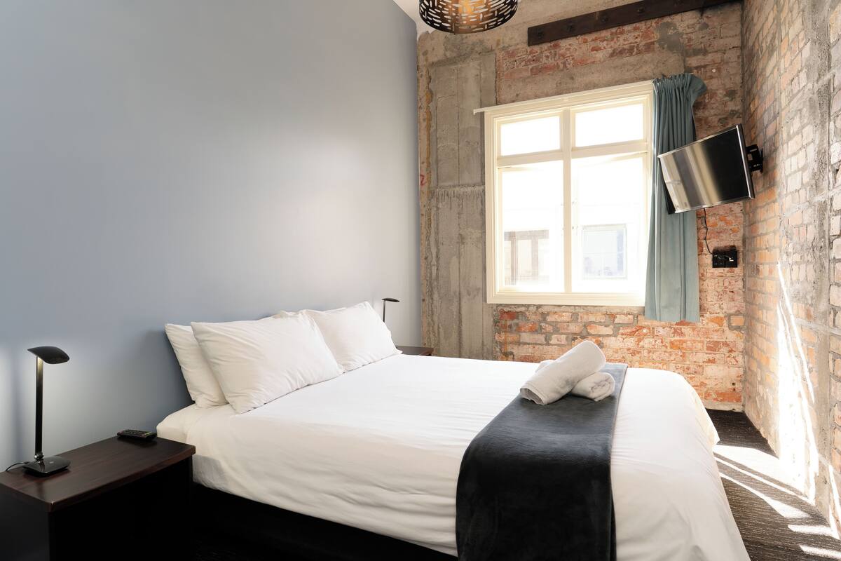 A neatly made bed with white linens, a black throw blanket, and a white towel folded at the foot. A black floor lamp is on a wooden nightstand beside the bed. A flat-screen TV is mounted on the brick wall to the right of the bed.