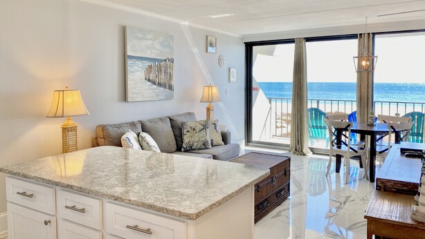 A modern hotel room with a large window offering a view of the sea, a cozy seating area with a table and chairs, and a marble island with a sink.