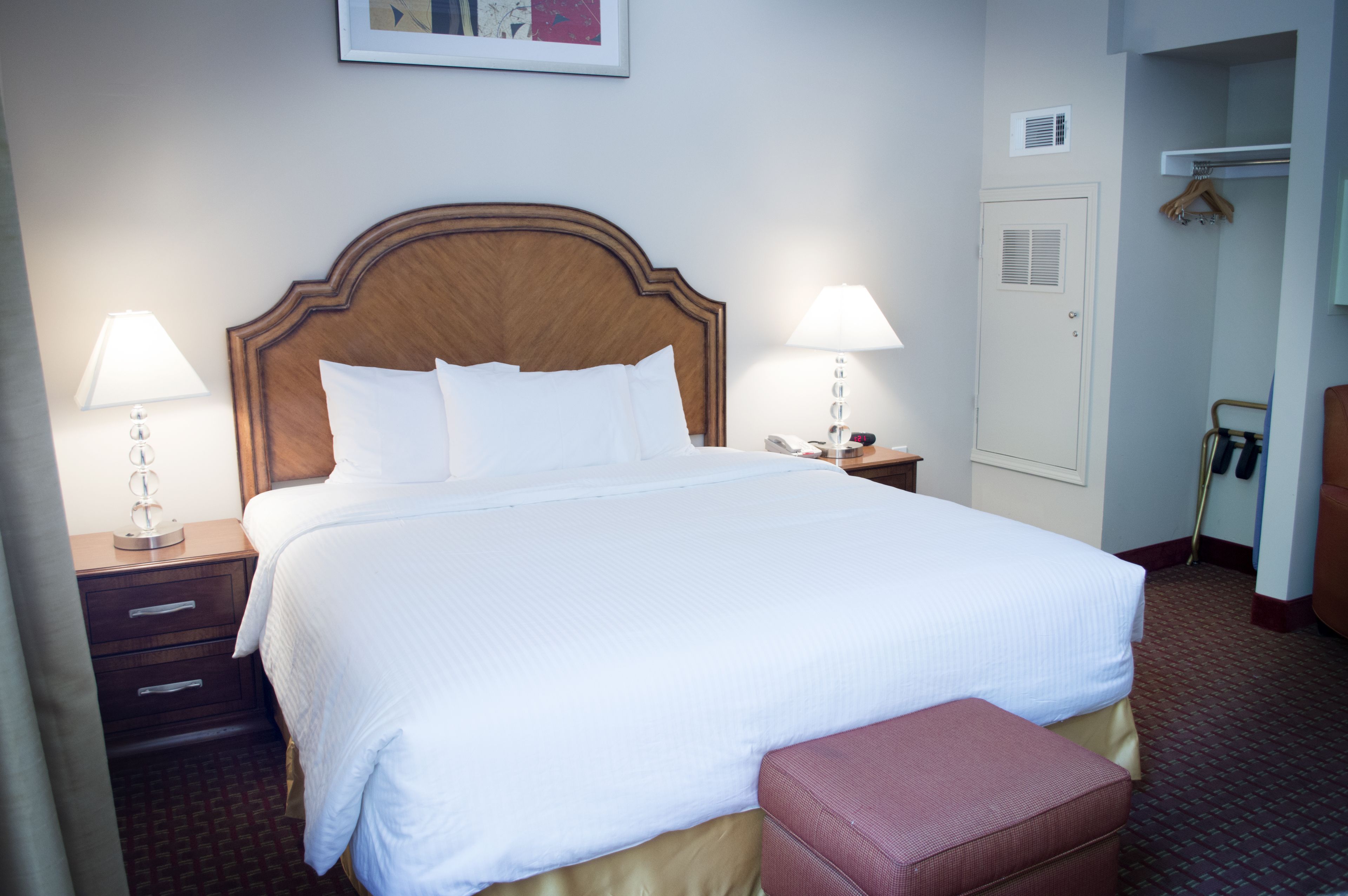 A hotel room with a large bed, two bedside tables with lamps, a wooden headboard, a wardrobe, and a small bench.