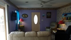 Uma sala de estar com sofá bege, uma cadeira, uma mesinha com abajur e uma porta com janela.