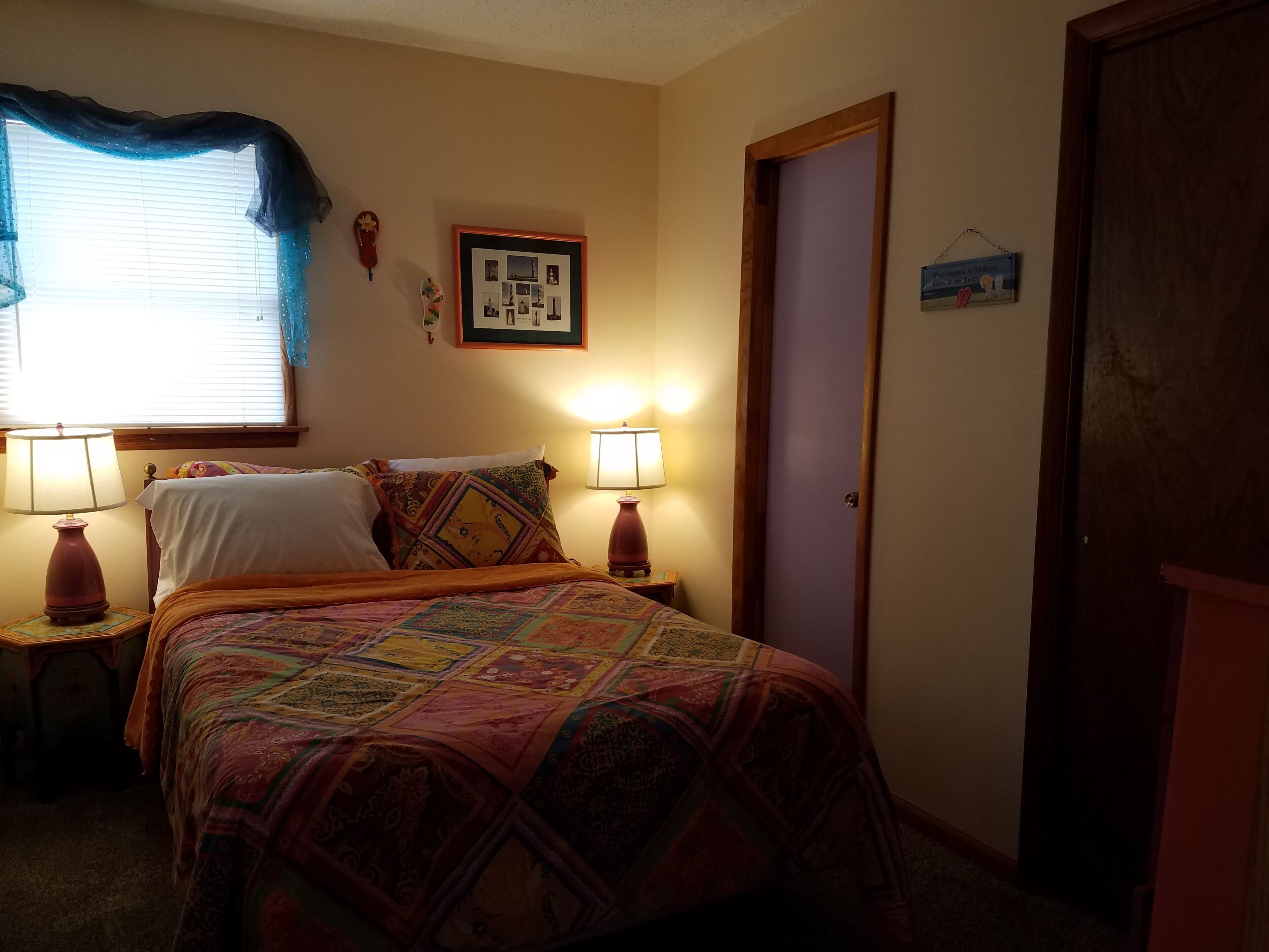 A bedroom with a bed, two bedside lamps, a framed photo, a door, and a window with a blue curtain.