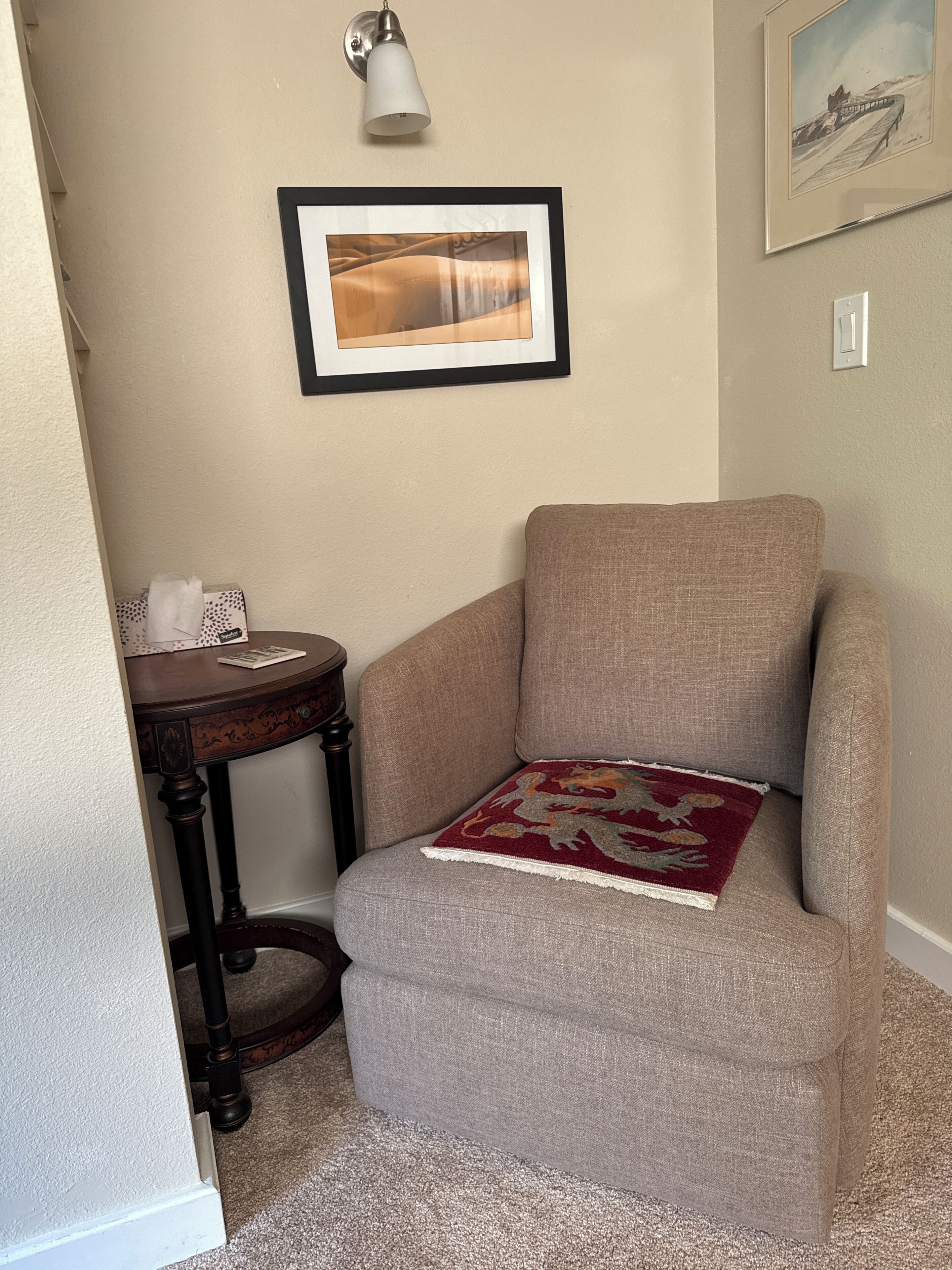 A corner room with a beige armchair, a small table with a lamp, and a framed picture on the wall.