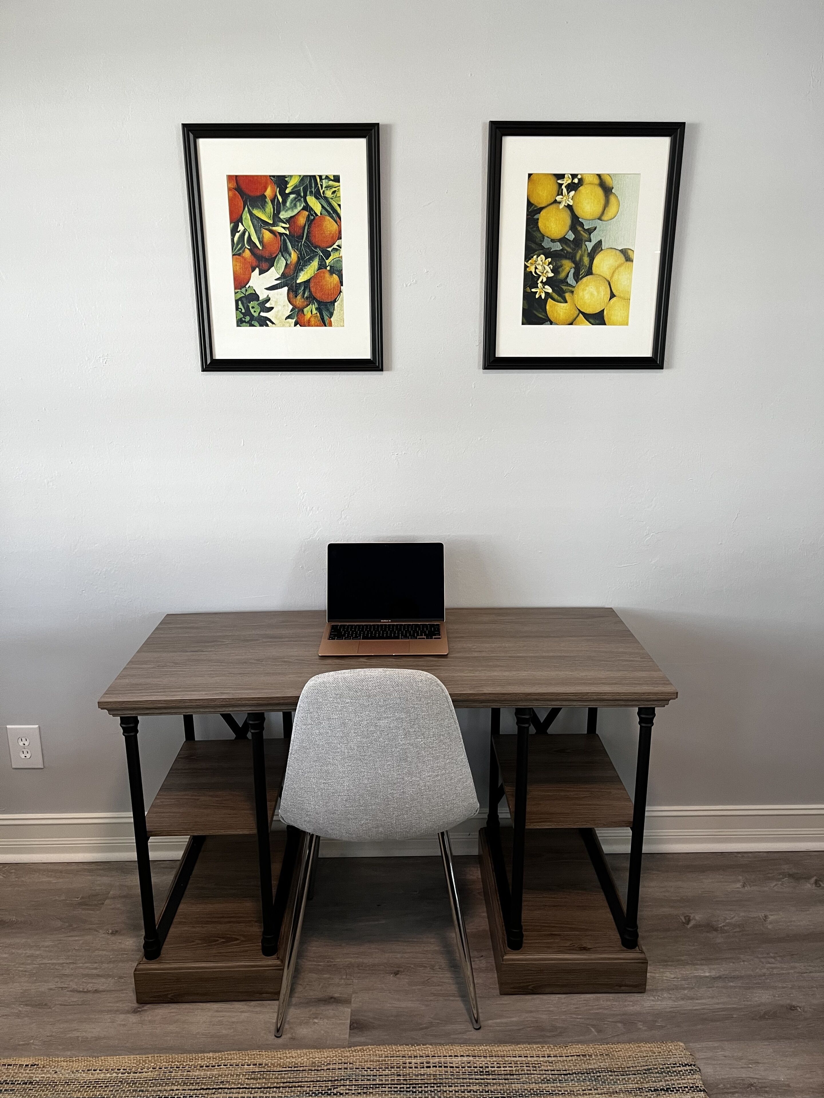 Un espace de travail avec un ordinateur portable posé sur une table en bois, une chaise grise et deux œuvres d’art encadrées accrochées au mur.