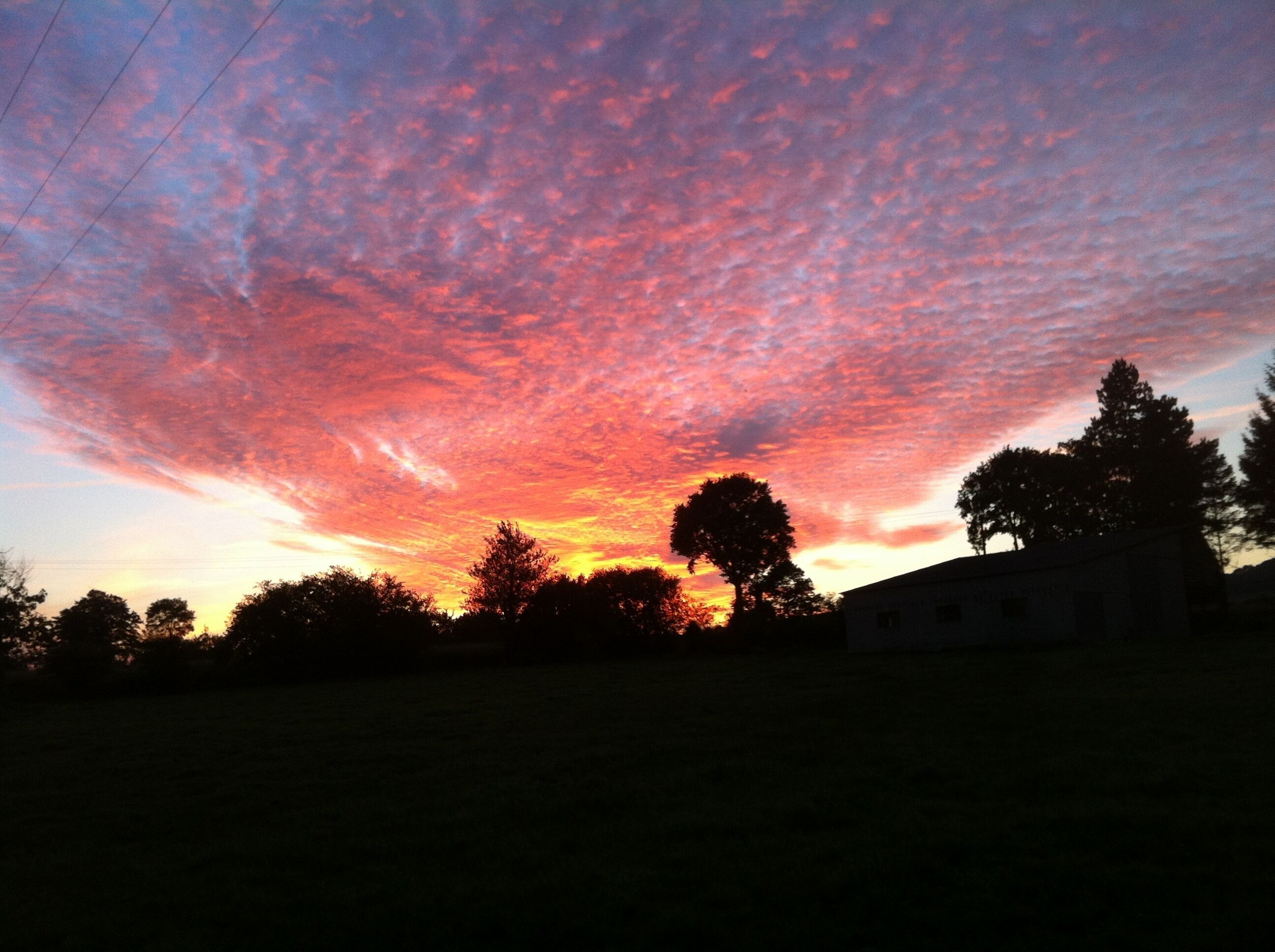 Ein Sonnenuntergang mit einem leuchtenden Himmel, Baumkonturen und einem Gebäude im Vordergrund.