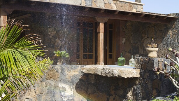 Un edificio de piedra con una cascada, un banco de piedra y una palmera.