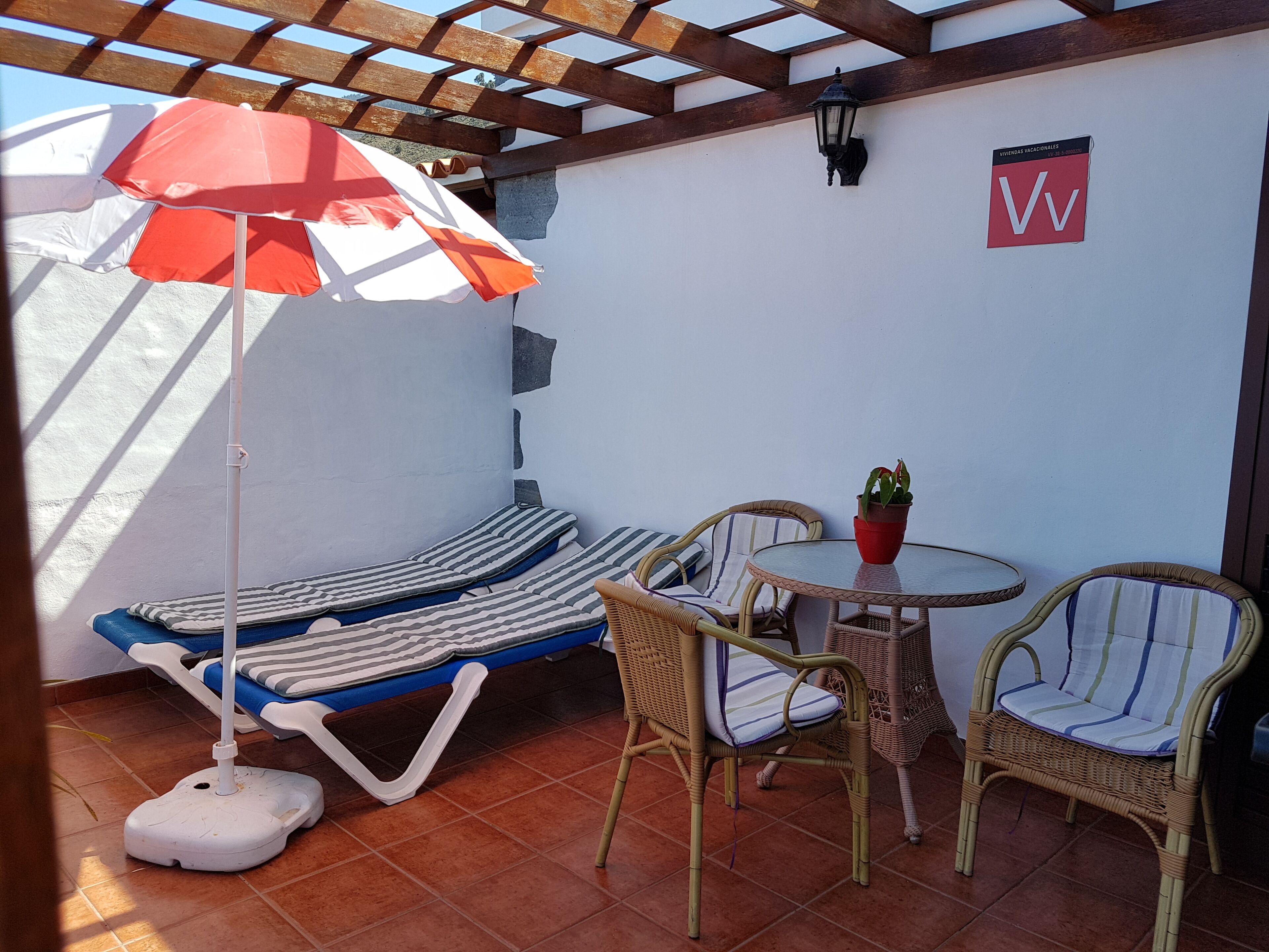 Un patio soleado con una silla de salón, cojines a rayas, una mesita con una planta en maceta y sillones de mimbre.