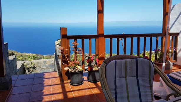 Un balcon en bois offrant une vue sur la mer, agrémenté de plantes en pot et d’une chaise à rayures.