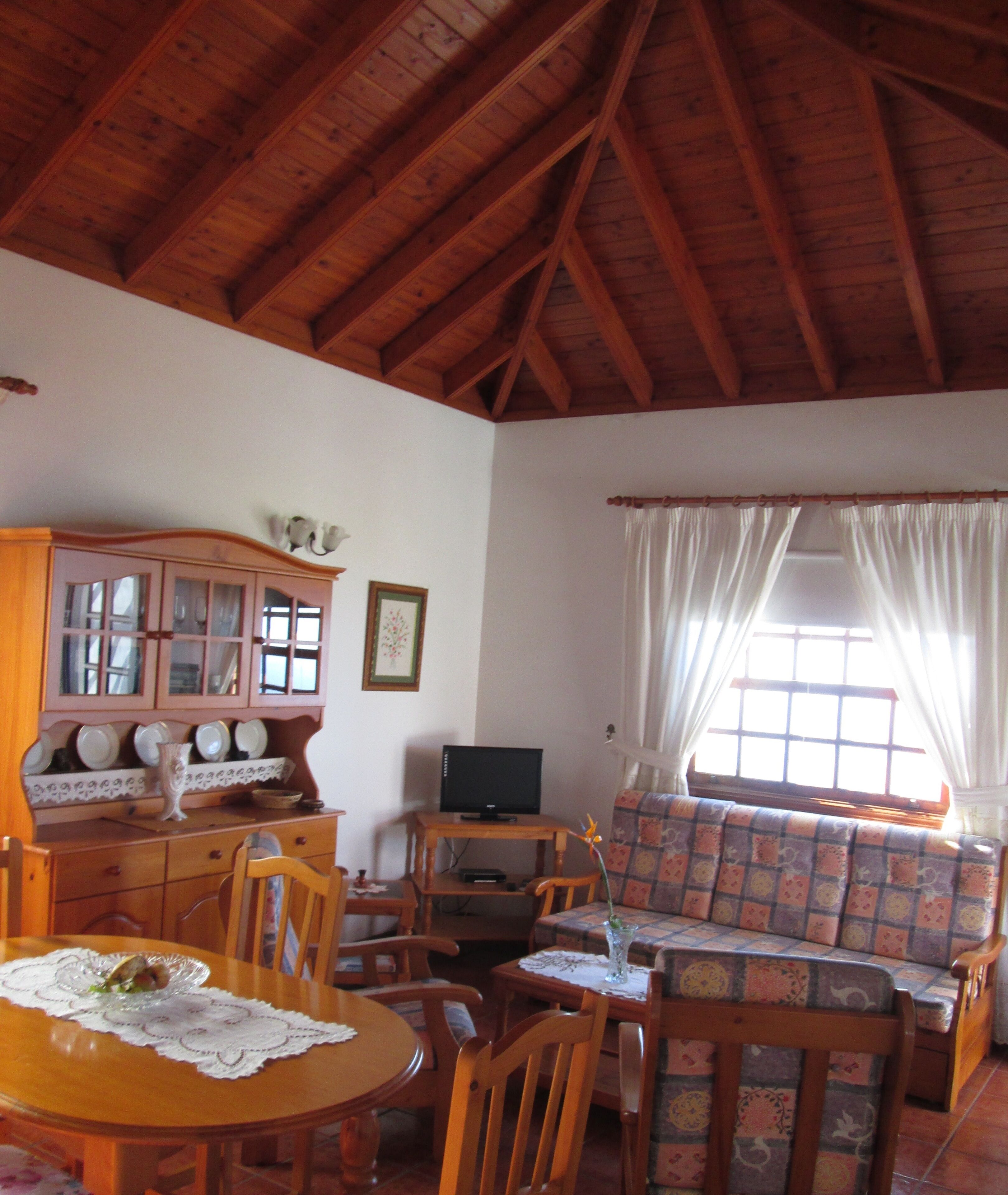 A cozy living room with wooden beams, a dining table, chairs, a sofa, and a television.