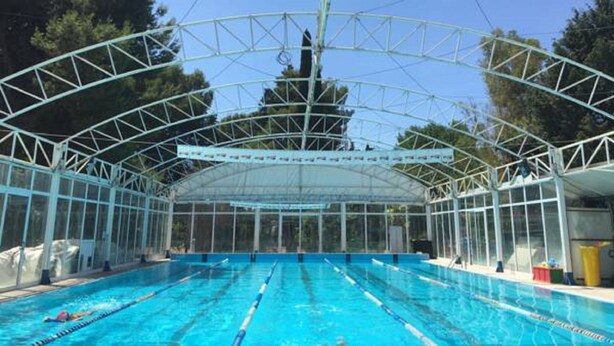Une piscine couverte avec des couloirs bien délimités, une structure de toit en métal et une vue sur les arbres en arrière-plan.