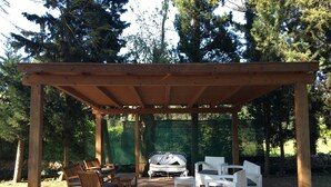 Une pergola en bois avec un espace salon extérieur comprenant des chaises et une table, entourée de plantes en pot et d’un barbecue.
