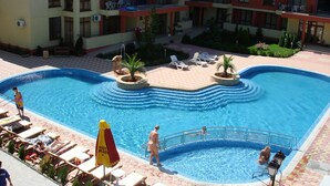 Una piscina con sillones de descanso y sombrillas, rodeada por un edificio de varios pisos con balcones.
