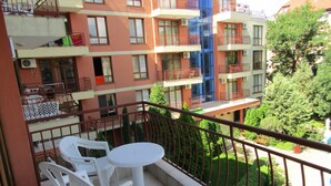 Un balcón con sillas de plástico blanco y una pequeña mesa redonda, con vistas a un jardín y edificios contiguos con balcones.