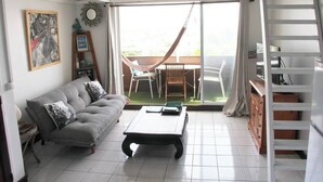 A living room with a sofa, a coffee table, a dining table with chairs, a hammock, and a sliding glass door leading to an outdoor area.