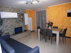 A modern living room with a stone wall, a TV, a dining table with chairs, and a vase with flowers.