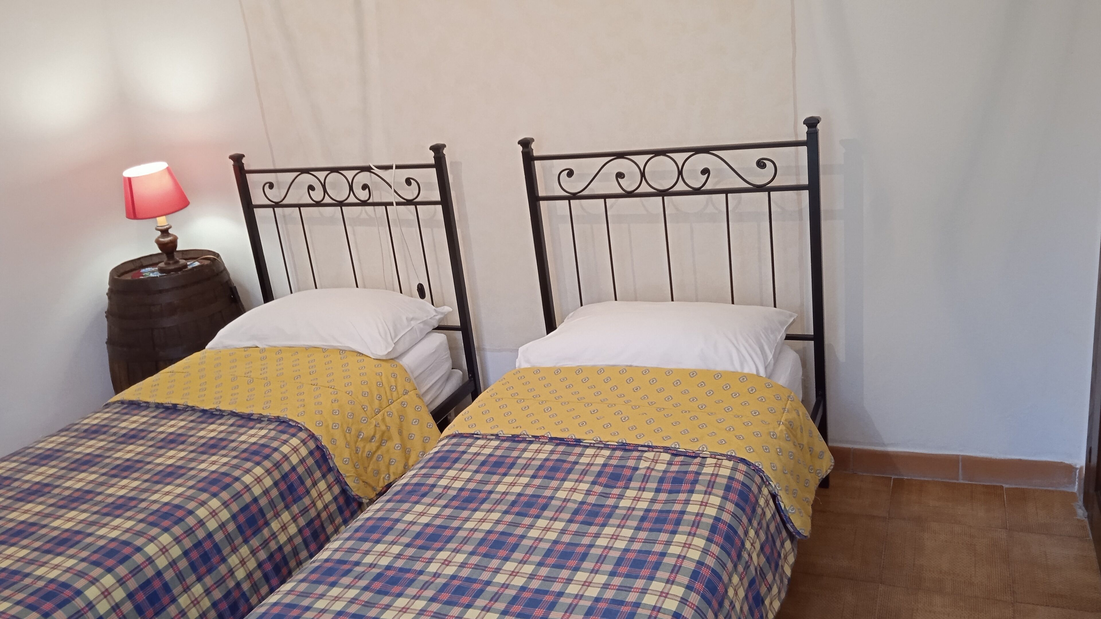 A room with two beds, each with a patterned bedspread and pillows, a nightstand with a lamp, and a wooden barrel.