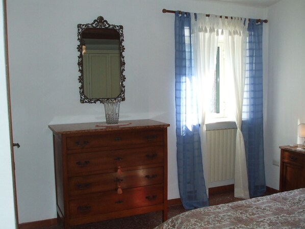 Ein Schlafzimmer mit einem Holzkästchen, einem Spiegel, einem Fenster mit blauen Vorhängen und einem Nachttisch mit einer Lampe.
