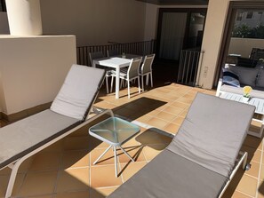 Un espacio al aire libre iluminado por el sol, con dos sillones de descanso y una pequeña mesa, rodeado de un suelo de baldosas y una barandilla de metal.