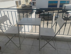 Un balcon avec une table blanche et deux chaises blanches, une rambarde en métal, et une vue sur les voitures garées et les immeubles.