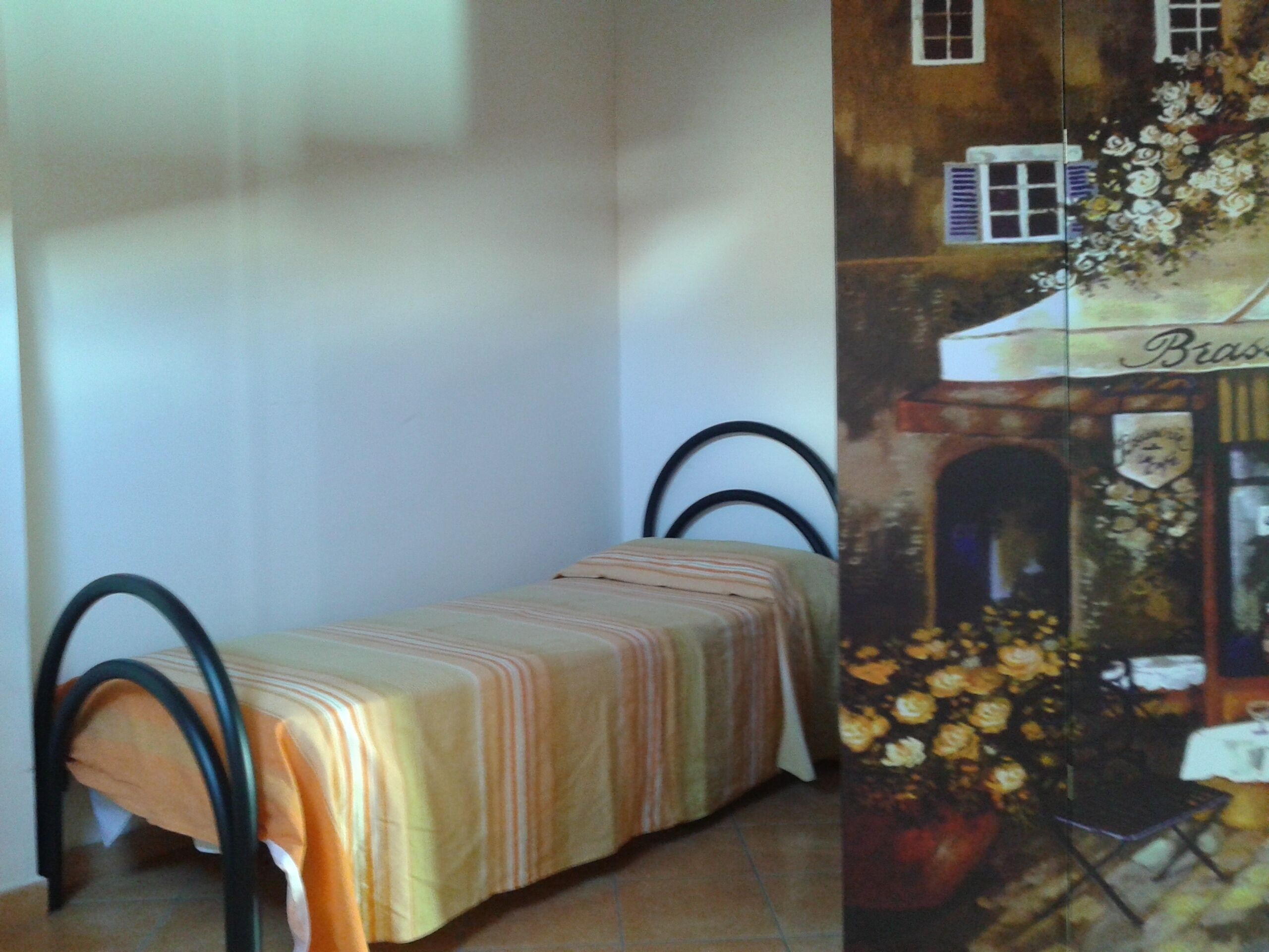 A small, single-bed room with a striped bedspread and a metal frame bed. There is a wall with a painting of a building and flowers.