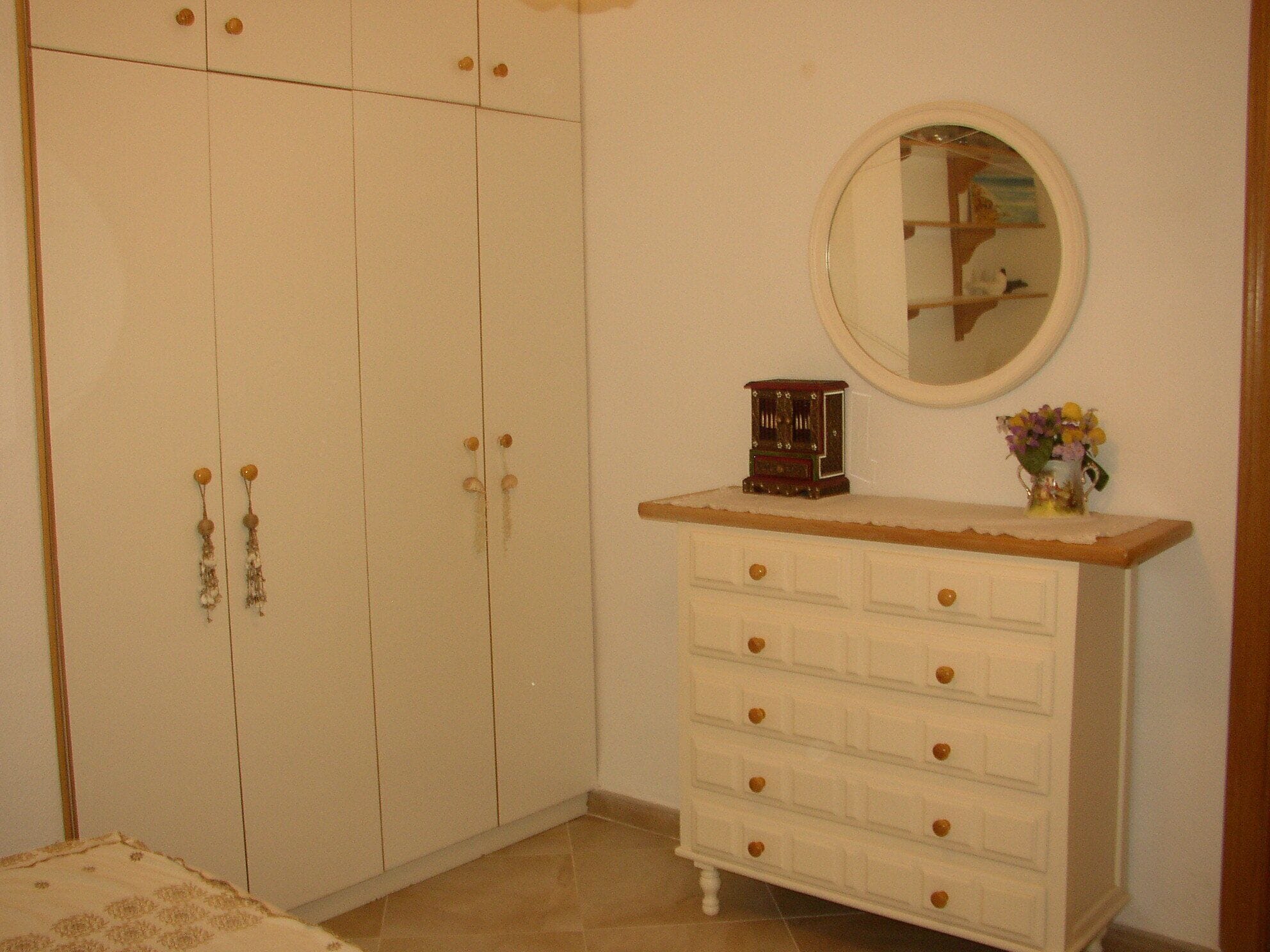 A room with a white wardrobe, a dresser with drawers, a mirror, and a small clock on the dresser.