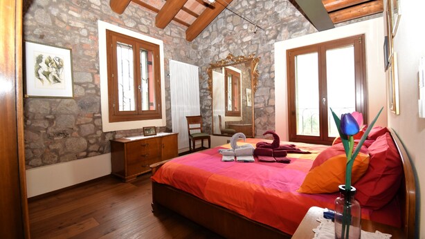 A bedroom with a stone wall, wooden ceiling beams, a bed with an orange bedspread, a wooden dresser, a chair, a mirror, and a window with a view.
