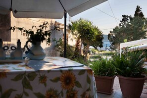 Un área de comedor al aire libre con una mesa cubierta por un mantel floral, plantas en macetas y vista al mar.