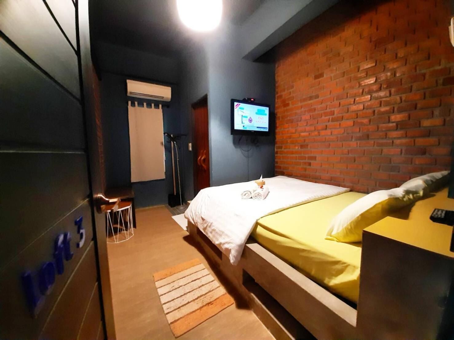 A hotel room with a bed, a TV, a brick wall, and a window.
