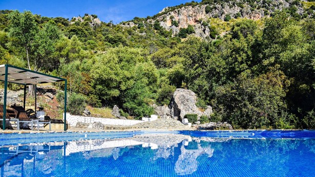 Une piscine d’un bleu éclatant, bordée d’une terrasse en pierre et dotée de chaises longues, le tout entouré d’une végétation luxuriante et de collines rocheuses.