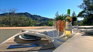 Une table dressée pour deux personnes, avec une bouteille de vin, des verres et une assiette de nourriture, donnant sur un paysage montagneux.