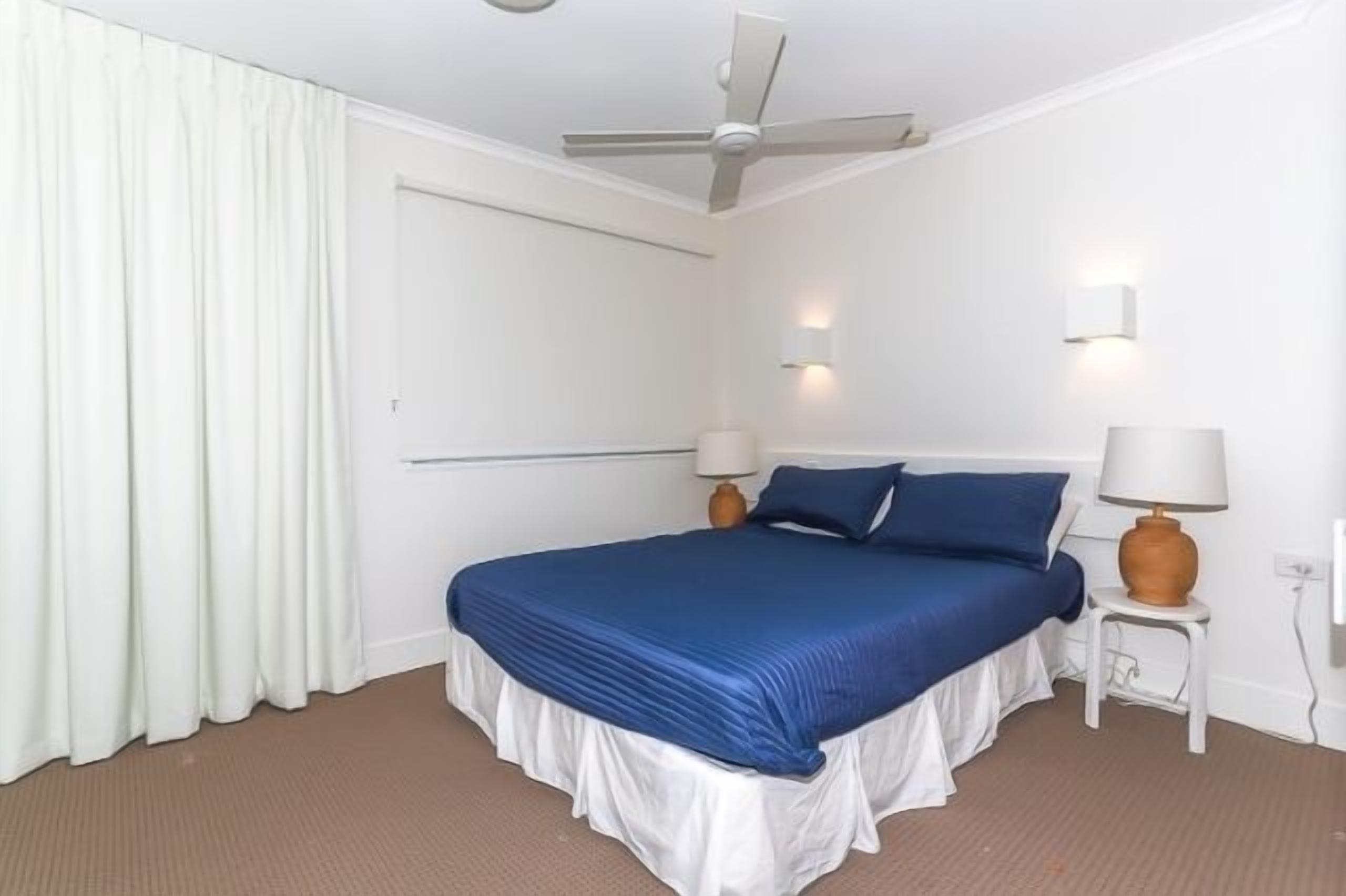 A hotel room with a bed, two bedside lamps, a ceiling fan, and a window with curtains.
