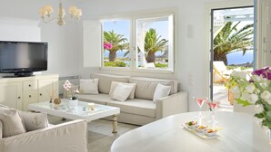 Een moderne woonkamer met een bank, een salontafel en een televisie. Er zijn ramen met uitzicht op palmbomen en de zee.