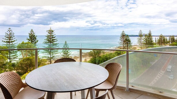 A balcony with a round table and chairs, overlooking a beach and ocean. - Breeze Apartment 801 (Mooloolaba)