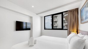 A modern hotel room with a large bed, a flat-screen TV mounted on the wall, a window with a view of a building, and a bedside table with a lamp. - Breeze Apartment 801 (Mooloolaba)